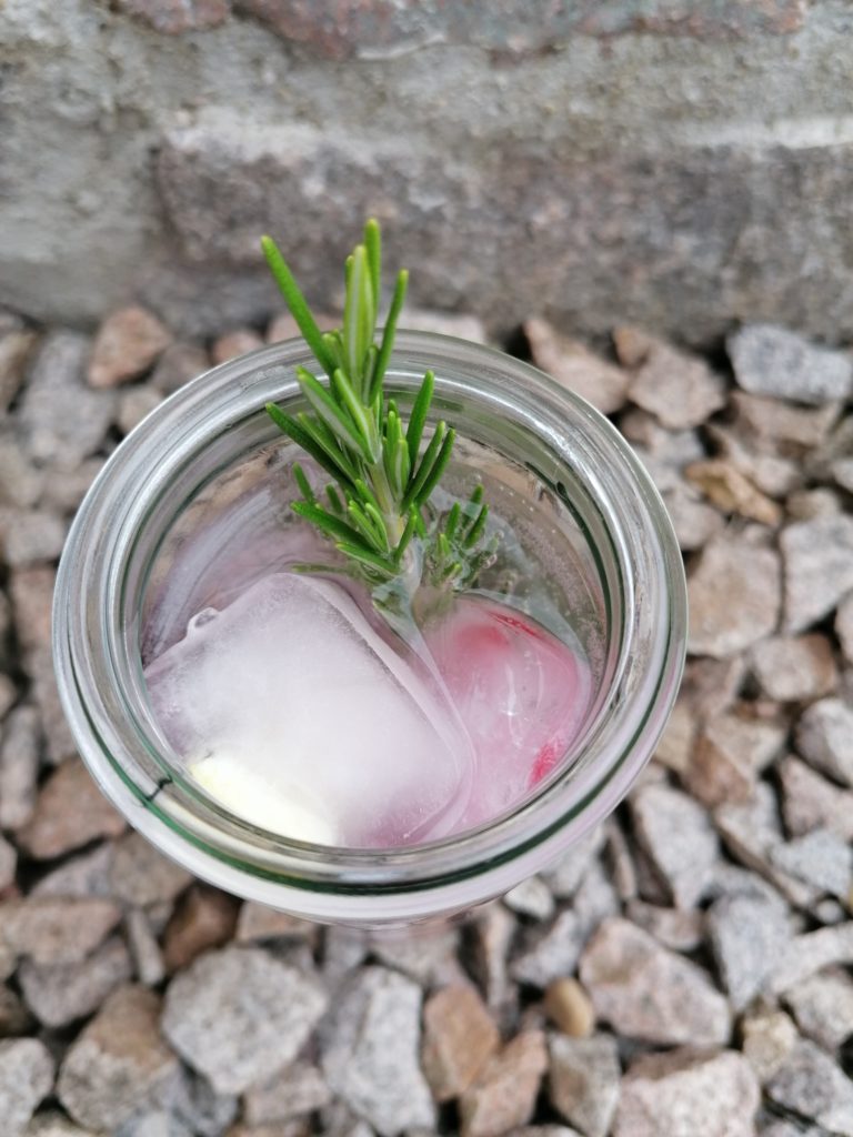 Infused Water mit Rosmarin und Eiswürfel von oben fotografiert. Das Glas steht auf Steinen vor einer Steinmauer.