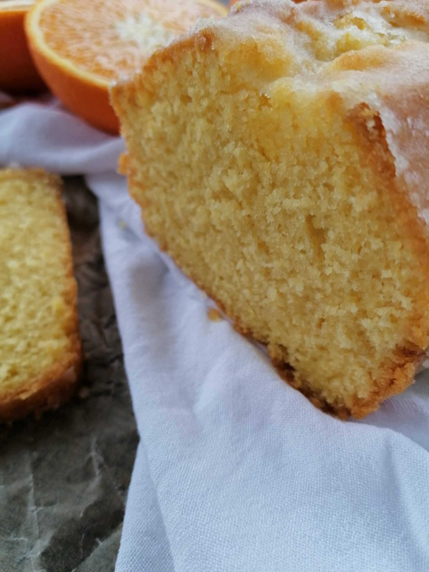 Orangenkuchen aufgeschnitten auf einem weißen Tuch. Im Hintergrund eine halbe Orange. Links im Bild liegt ein abgeschnittenes Kuchenstück.